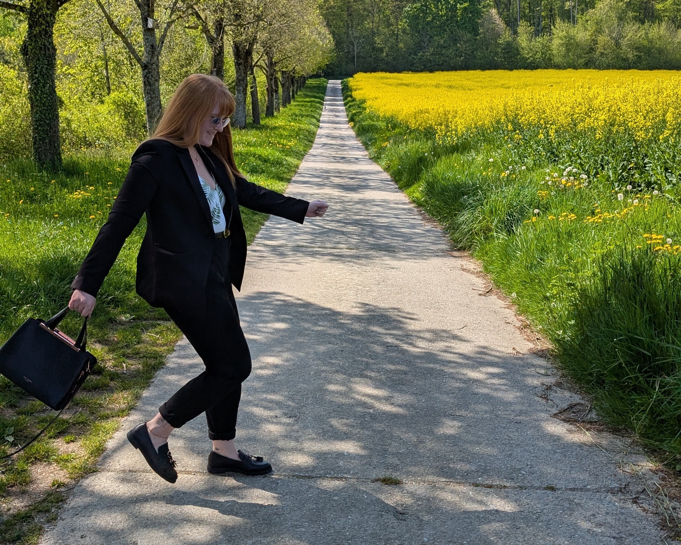 Une jeune femme rousse en costume noir, se promenant dans un champ de colza, Mathilda, coach en marketing Digital et Human Design